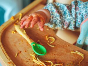 En barnehånd griber ud efter en ske på en højstol fyldt med spaghetti